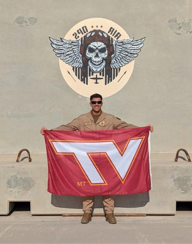U.S. Navy Lt. 古普塔拿着弗吉尼亚理工大学的旗帜微笑着. 他穿着米色飞行服，戴着墨镜，站在写着“空中行动”的彩绘艺术品下面.”